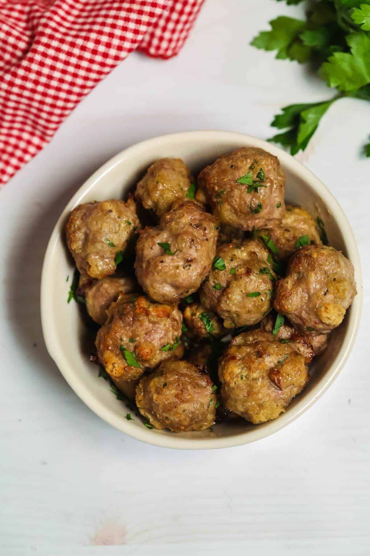 A bowl of baked meatballs garnished with chopped herbs sits on a white surface, accompanied by a red checked cloth and vibrant green leafy vegetables.