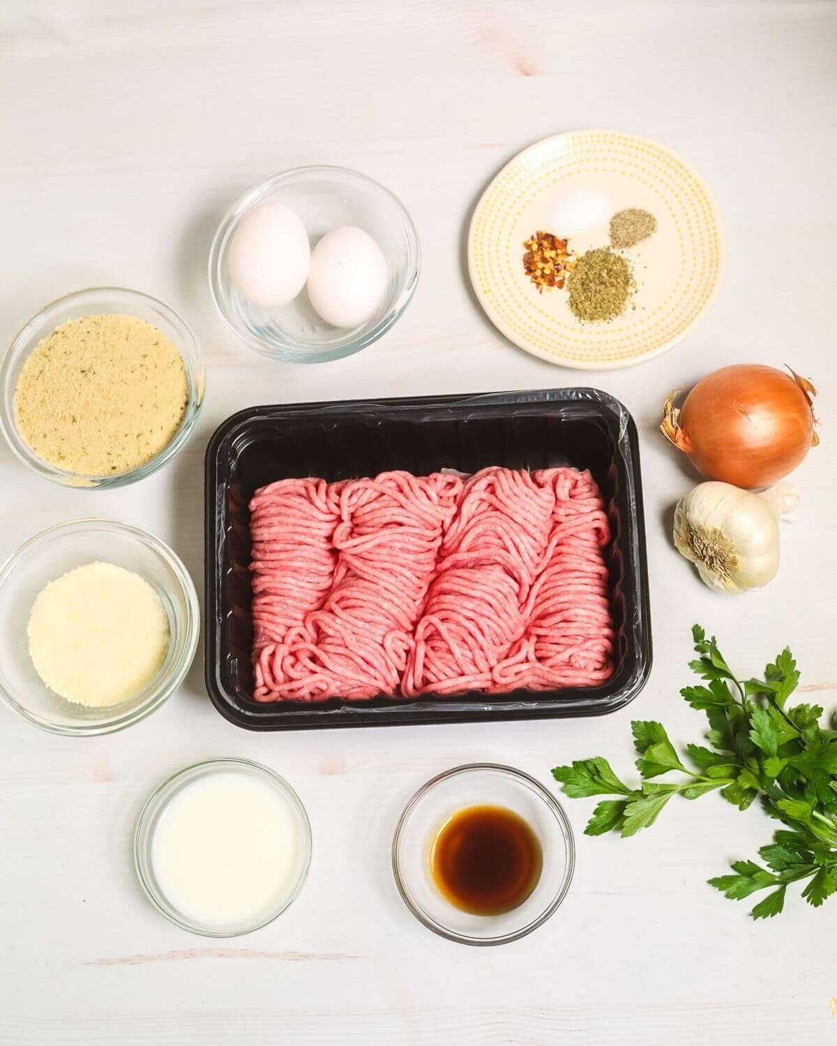 Ground meat nestled in a black tray, surrounded by fresh ingredients: two eggs, breadcrumbs, a seasoning mix, onion, garlic, milk, parmesan, Worcestershire sauce, and parsley on a light surface.