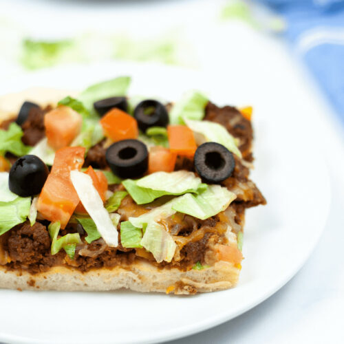 A slice of Mexican taco pizza topped with ground meat, lettuce, tomatoes, and black olives on a white plate.
