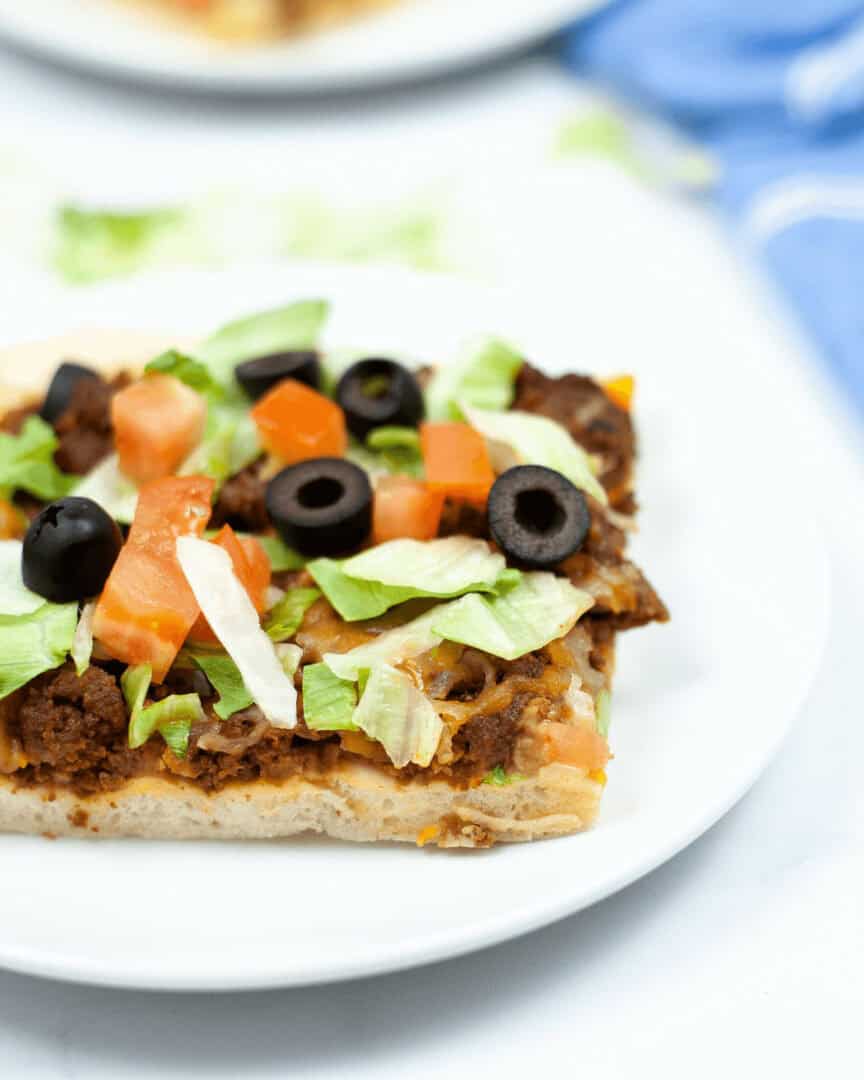A slice of Mexican taco pizza topped with ground meat, lettuce, tomatoes, and black olives on a white plate.