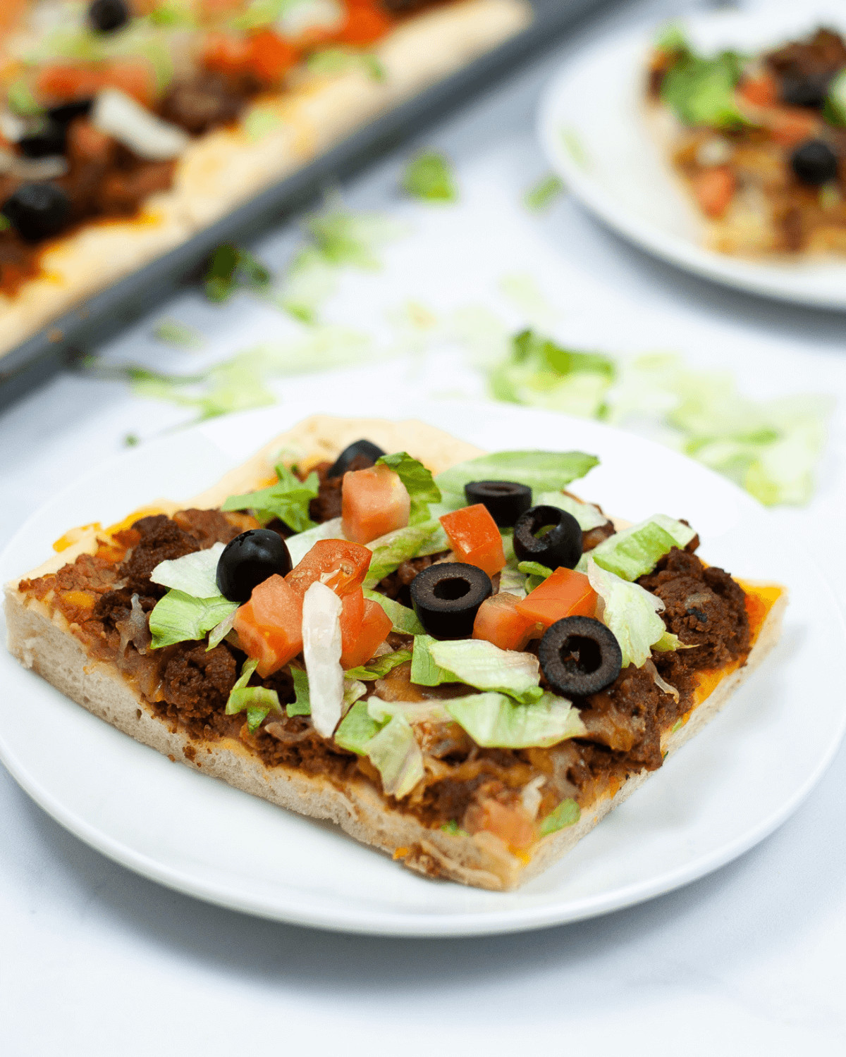 A slice with ground beef, lettuce, diced tomatoes, and black olives on a white plate, with more pizza in the background.