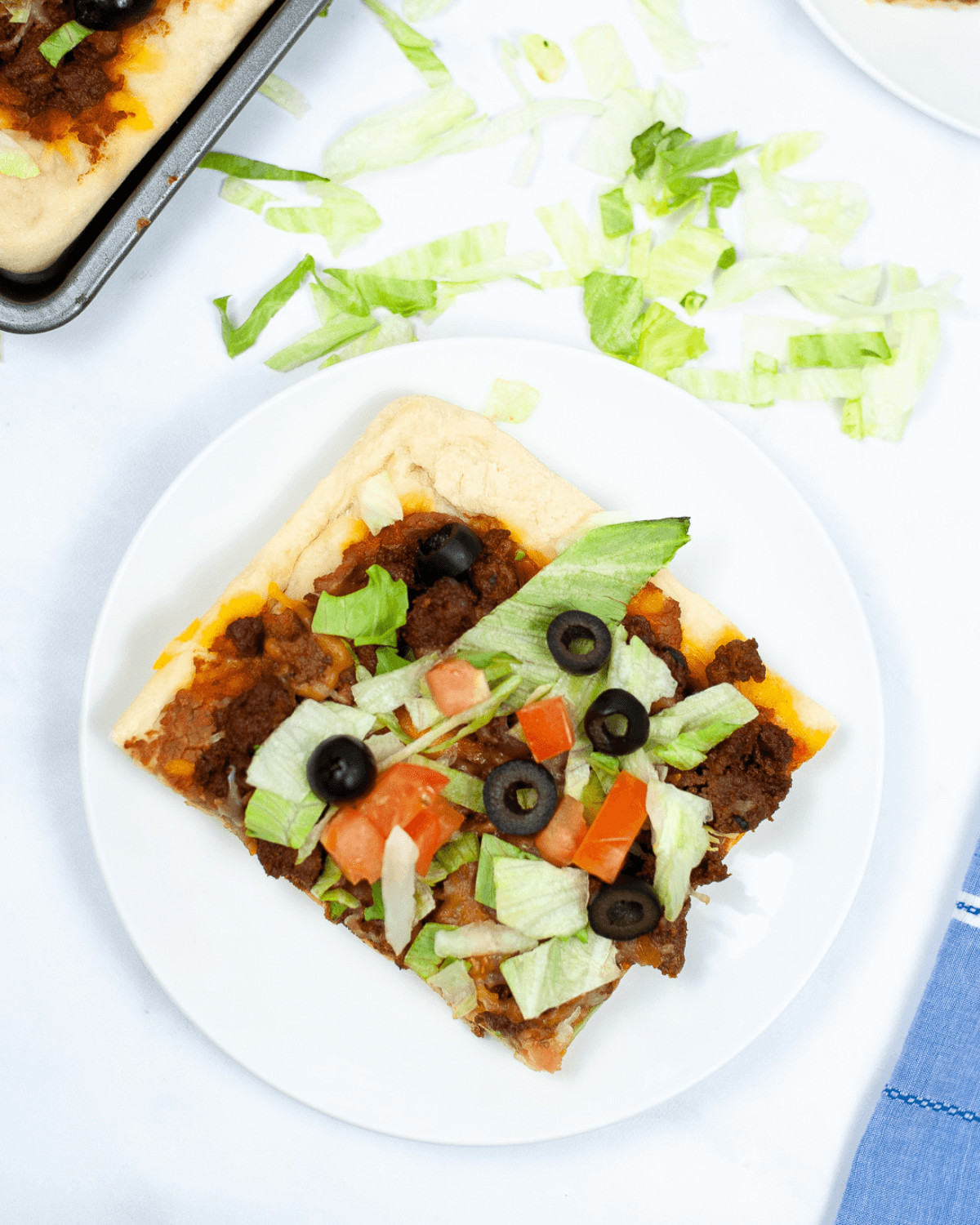 A slice of Mexican taco pizza topped with lettuce, tomatoes, and black olives on a white plate, with more pizza and lettuce in the background.