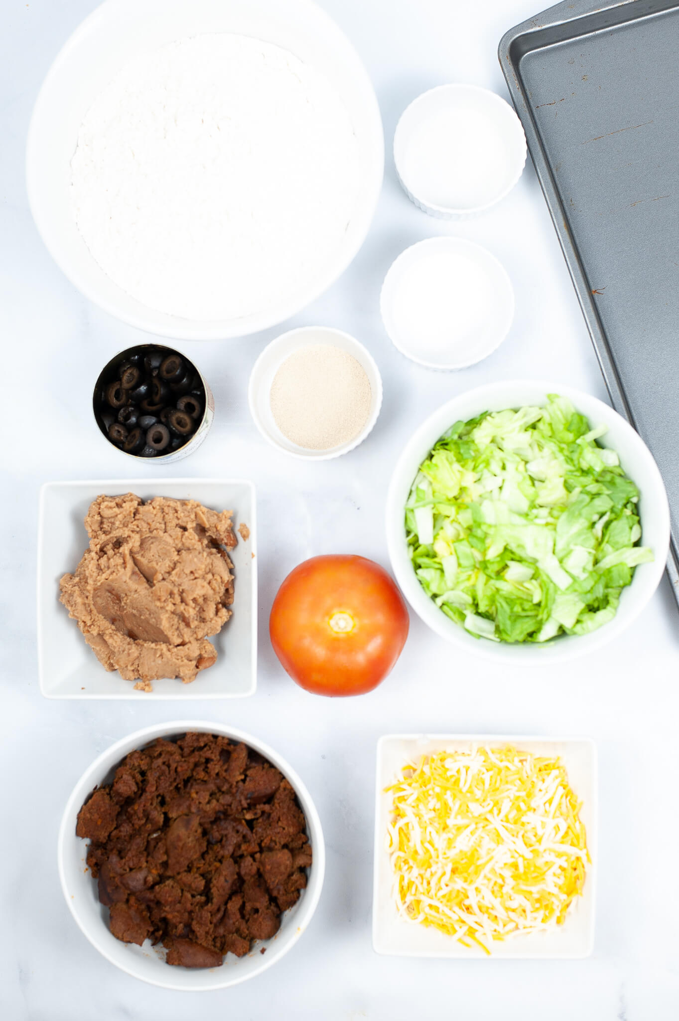 Ingredients for a Mexican pizza recipe laid out on a table, including flour, olives, yeast, lettuce, refried beans, tomato, ground meat, and shredded cheese.