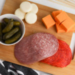 A wooden cutting board with meats, cheeses and pickles.