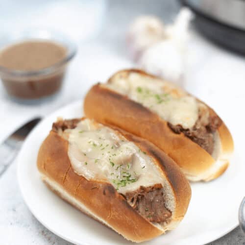 Two beef sandwiches with melted cheese and herbs sit enticingly on a white plate, echoing the flavors of a classic Instant Pot French Dip. They're accompanied by a savory dipping sauce, with a hint of garlic bulb in the background adding to the aromatic experience.