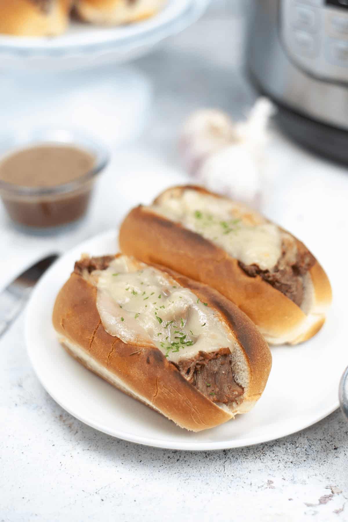 Two beef sandwiches with melted cheese and herbs sit enticingly on a white plate, echoing the flavors of a classic Instant Pot French Dip. They're accompanied by a savory dipping sauce, with a hint of garlic bulb in the background adding to the aromatic experience.