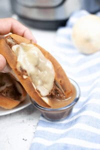 A hand holds a meal with melted cheese, dipping it into a small bowl of sauce, creating the perfect Instant Pot French Dip moment. A striped blue and white cloth rests on the table, adding to the cozy atmosphere.