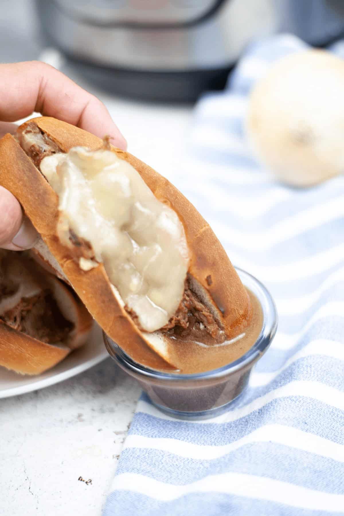 A hand holds a meal with melted cheese, dipping it into a small bowl of sauce, creating the perfect Instant Pot French Dip moment. A striped blue and white cloth rests on the table, adding to the cozy atmosphere.