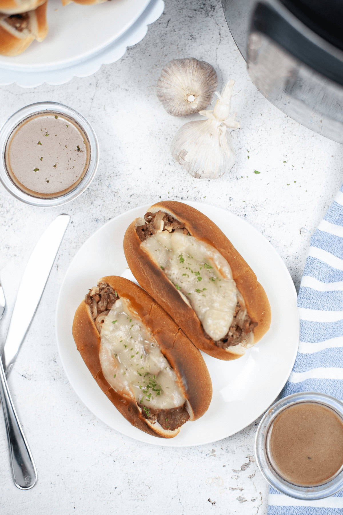 Two sandwiches with melted cheese and meat rest on a plate, surrounded by bowls of savory gravy, garlic cloves, and a striped napkin.