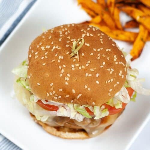 A sesame seed hamburger with lettuce, tomato, and cheese on a white plate, served with a side of sweet potato fries—try our Instant Pot Teriyaki Chicken Sandwich for a flavorful twist.