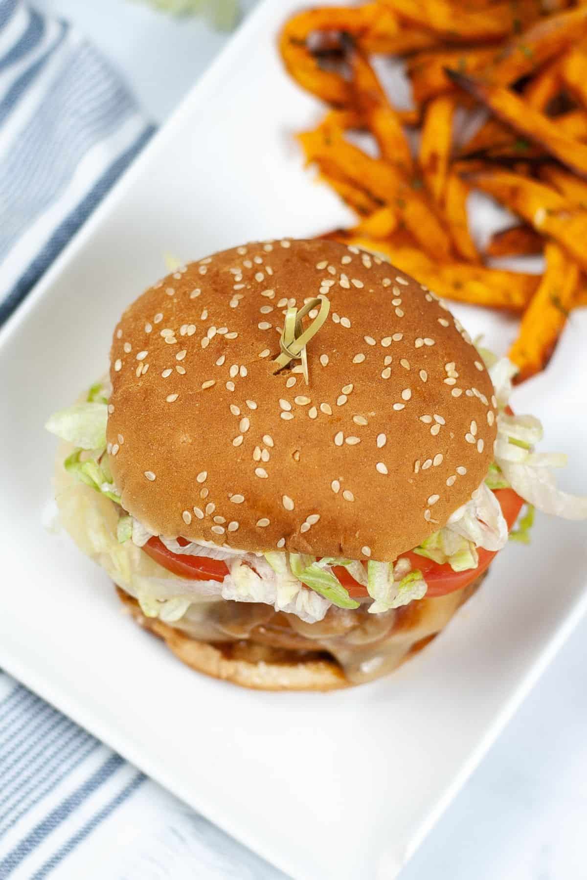 A sesame seed hamburger with lettuce, tomato, and cheese on a white plate, served with a side of sweet potato fries—try our Instant Pot Teriyaki Chicken Sandwich for a flavorful twist.