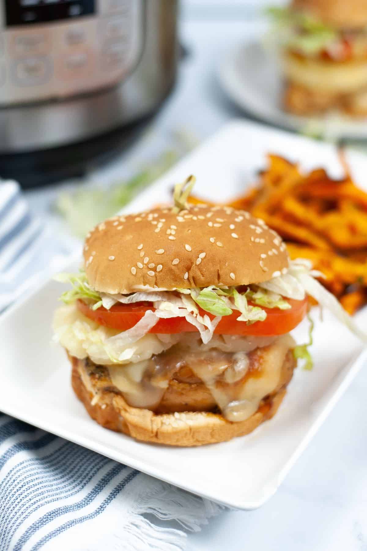 A cheeseburger with lettuce, tomato, and melted cheese on a sesame seed bun sits on a white plate next to a serving of sweet potato fries, sharing the spotlight with our flavorful Instant Pot Teriyaki Chicken Sandwich.