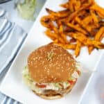 A sesame seed hamburger with lettuce and tomato is served on a white plate next to sweet potato fries, with fresh, vibrant toppings.