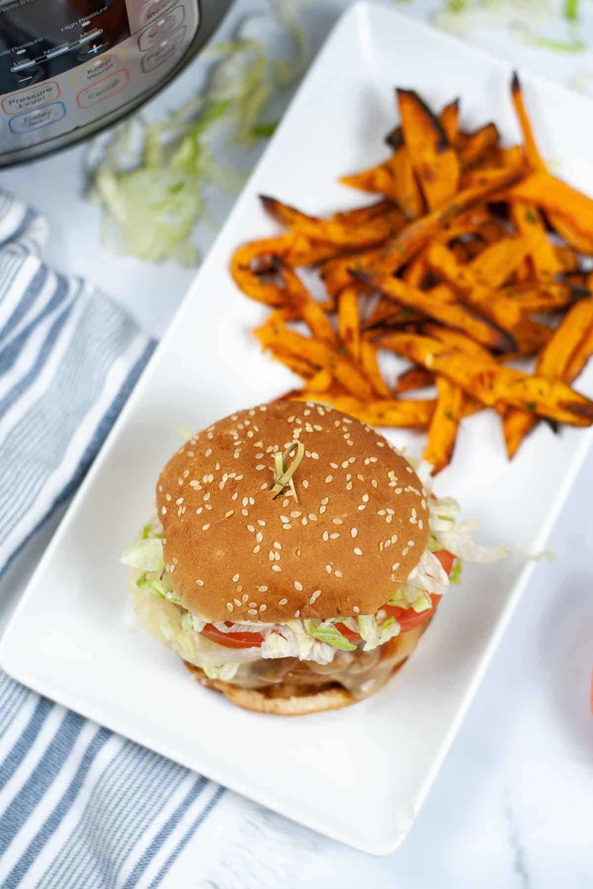 A sesame seed hamburger with lettuce and tomato is served on a white plate next to sweet potato fries, with fresh, vibrant toppings.