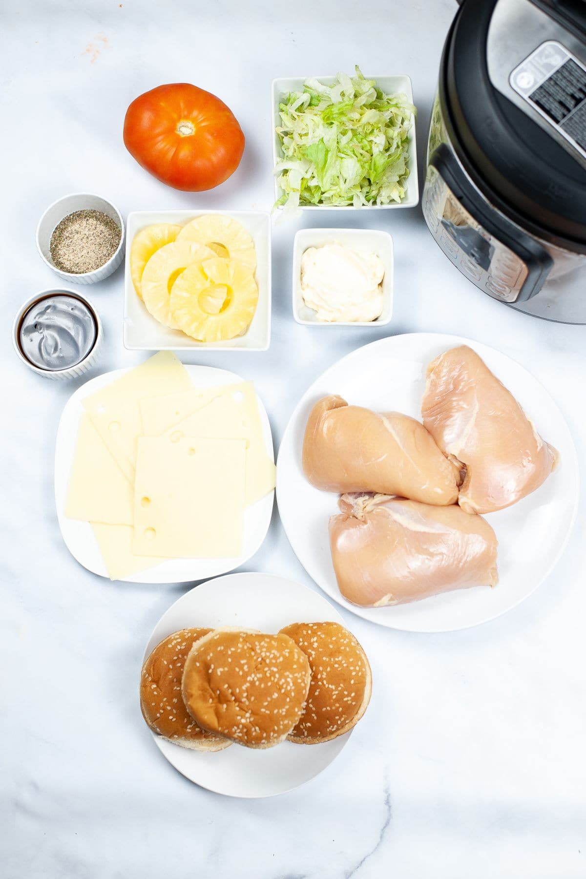 Ingredients arranged on a white surface, including raw chicken breasts, hamburger buns, cheese slices, lettuce, tomato, mayonnaise, seasonings.