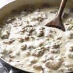 A skillet filled with creamy sausage gravy, perfect for topping fresh sausage biscuits, is being stirred with a wooden spoon.