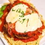 A serving of chicken parmesan or Veal Parmesan topped with melted cheese and marinara sauce, placed on a bed of spaghetti and garnished with chopped herbs.