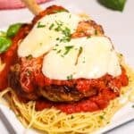 A plate of the dinner topped with melted cheese and tomato sauce, served over a bed of spaghetti and garnished with chopped herbs.