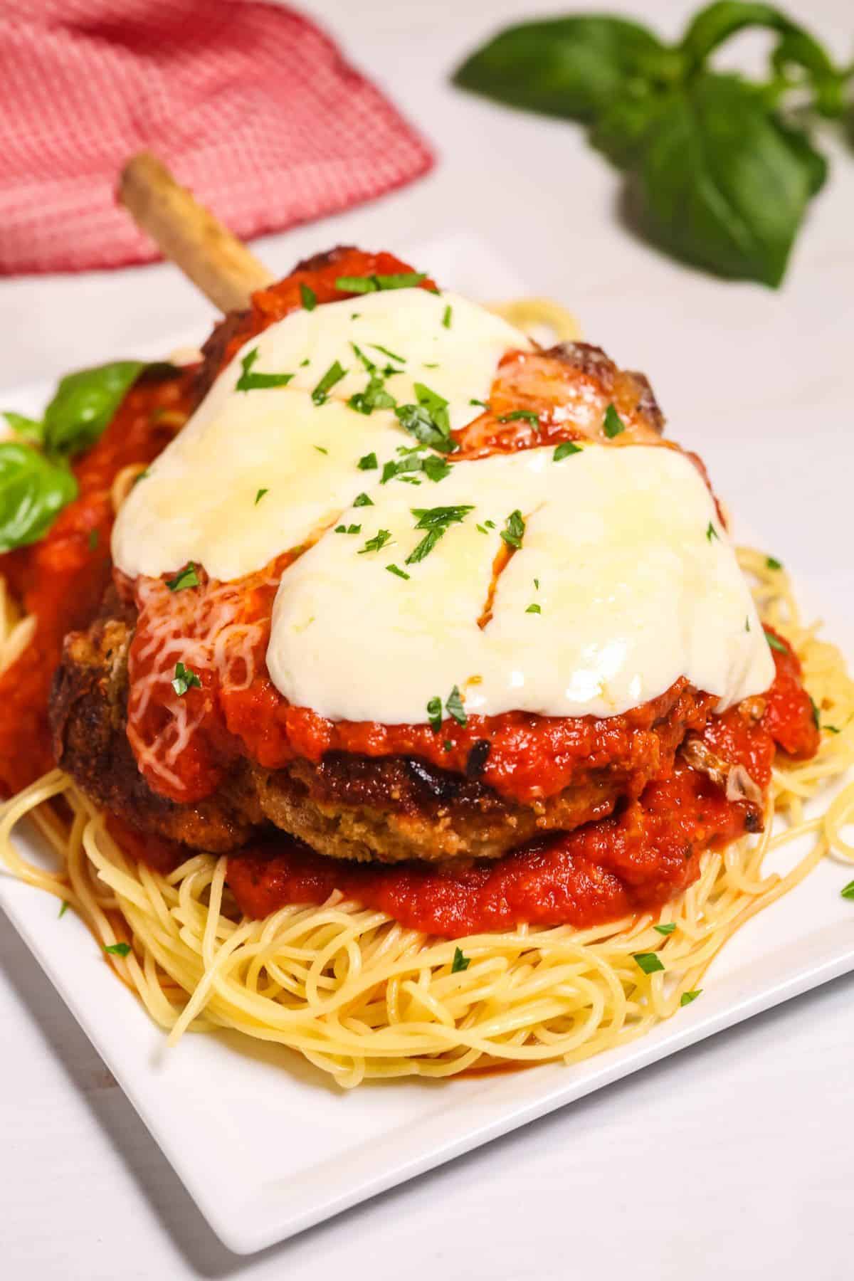 A plate of the dinner topped with melted cheese and tomato sauce, served over a bed of spaghetti and garnished with chopped herbs.