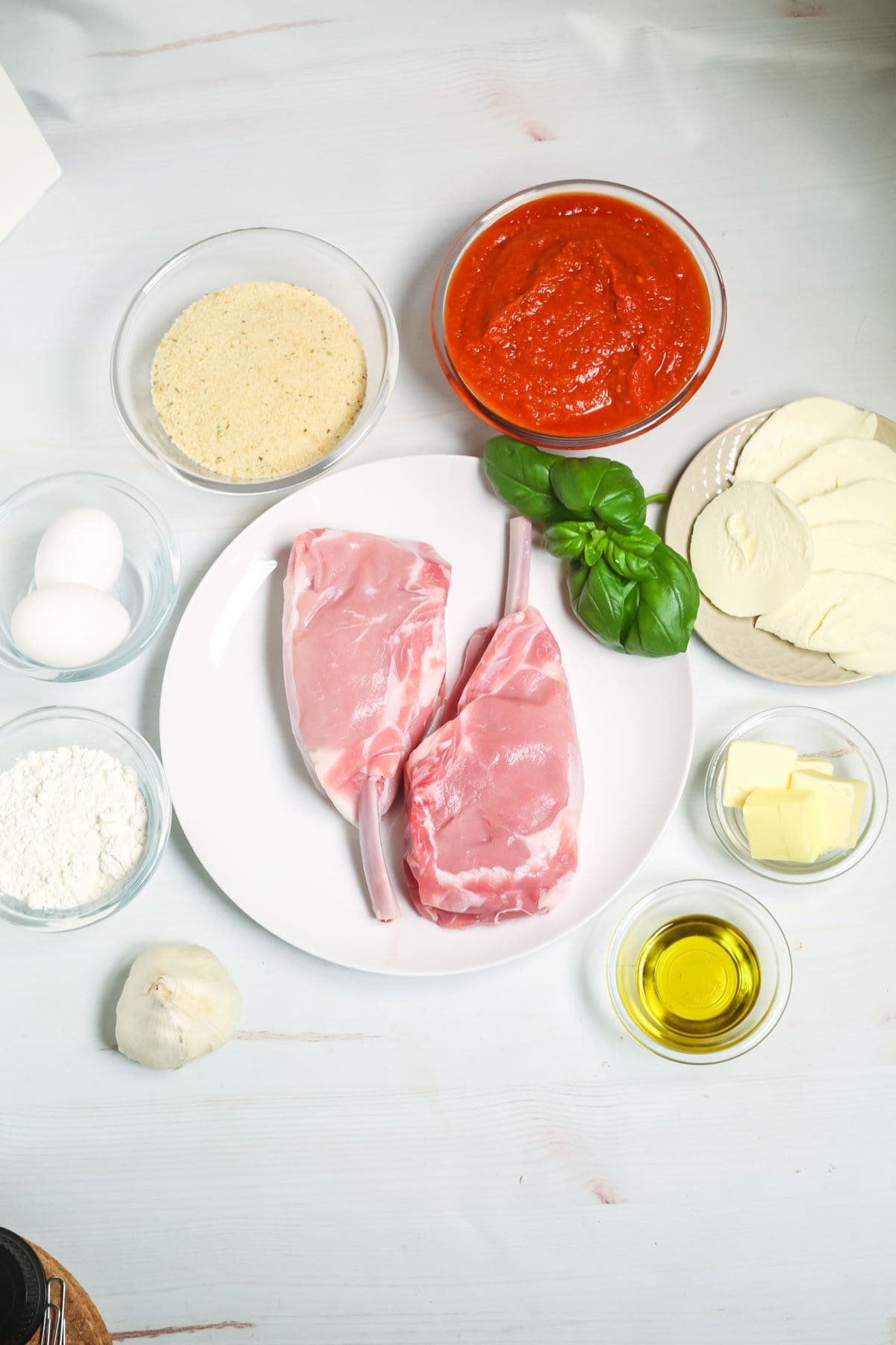 Two raw lamb chops on a plate surrounded by ingredients like eggs, flour, breadcrumbs, tomato sauce, mozzarella, and basil—perfect for creating classics such a experimenting with your own delicious twist.