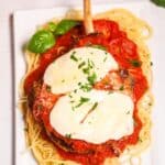 A classic Veal Parmesan: a breaded veal chop topped with melted mozzarella and tomato sauce, served over spaghetti, and garnished with basil and chopped parsley on a white rectangular plate.