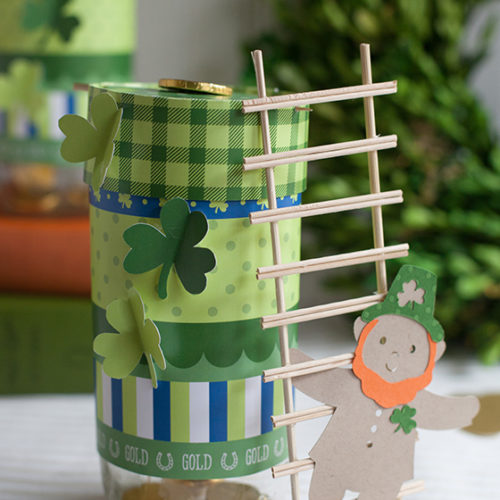 Tall glass jar decorate with shamrocks and green paper. A ladder leans against the festive jar.