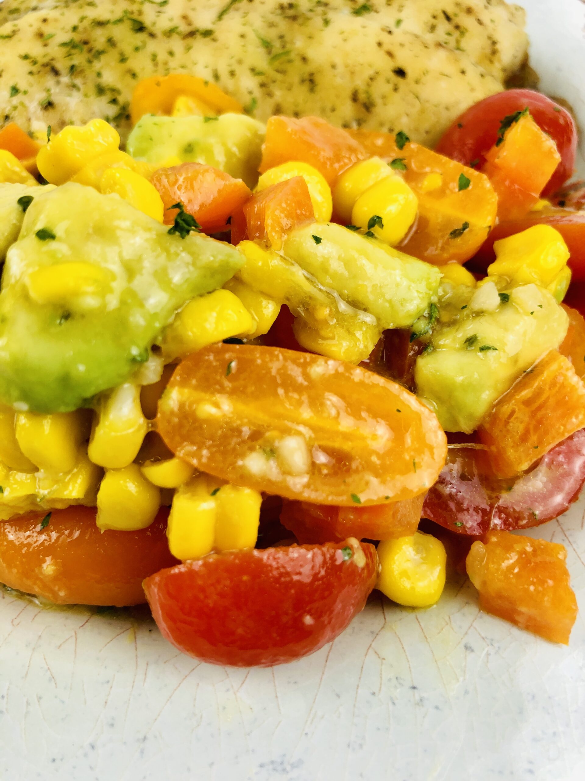 A colorful salad with cherry tomatoes, corn, and avocado dressing on a plate with baked haddock.
