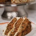 The Ultimate Carrot Cake on a white plate with a silver cake stand in the back.