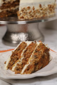 The Ultimate Carrot Cake on a white plate with a silver cake stand in the back.
