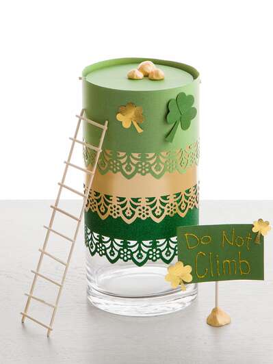 A glass jar decorated with multiple shades of green paper and shamrocks with a small wooden ladder leaning up against it.