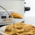 A forkful of pierogi over a plate with a crock pot in the background.