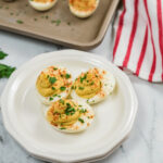Plate of Million Dollar Deviled Eggs garnished with paprika and parsley, with additional eggs and a striped napkin in the background.
