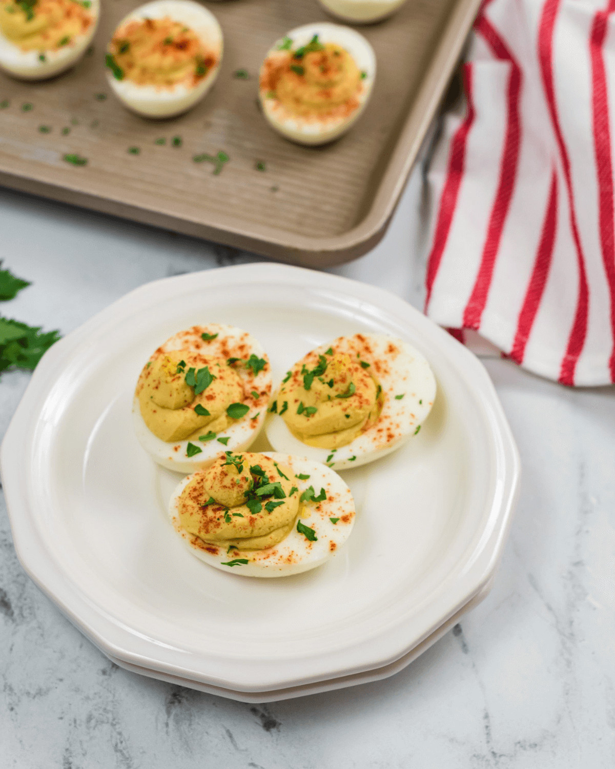 Plate of Million Dollar Deviled Eggs garnished with paprika and parsley, with additional eggs and a striped napkin in the background.