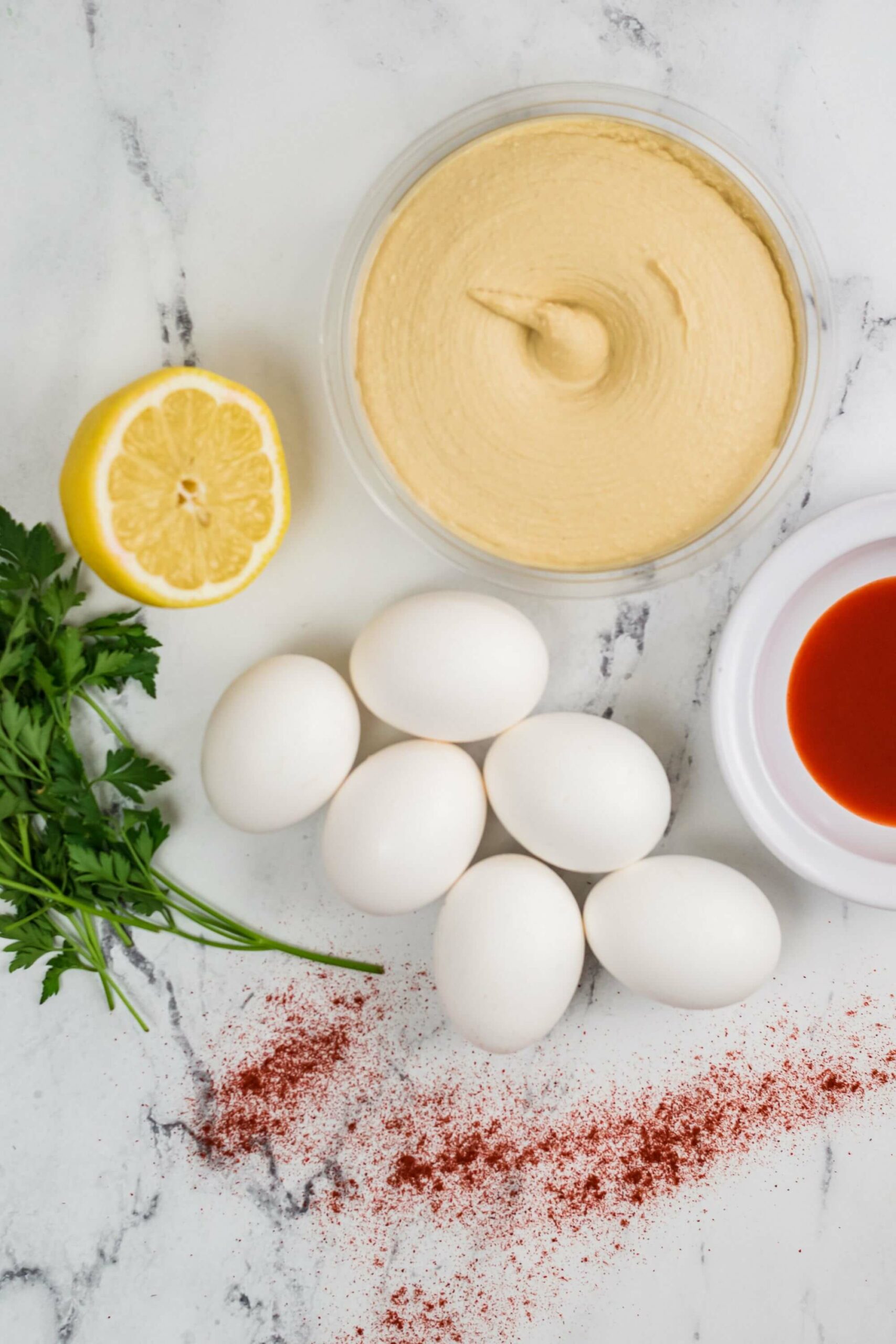 Ingredients for a Million Dollar Deviled Eggs recipe, including eggs, paprika, hot sauce, parsley, lemon, and a bowl of creamy sauce, on a marble surface.