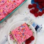 A slice of cake with raspberry buttercream frosting and rainbow sprinkles is served on a plate with raspberries; the rest of the cake and a basket of raspberries are nearby.