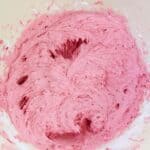 A bowl of vibrant pink raspberry buttercream is being mixed, with streaks of the creamy mixture and some flour visible around the edges.