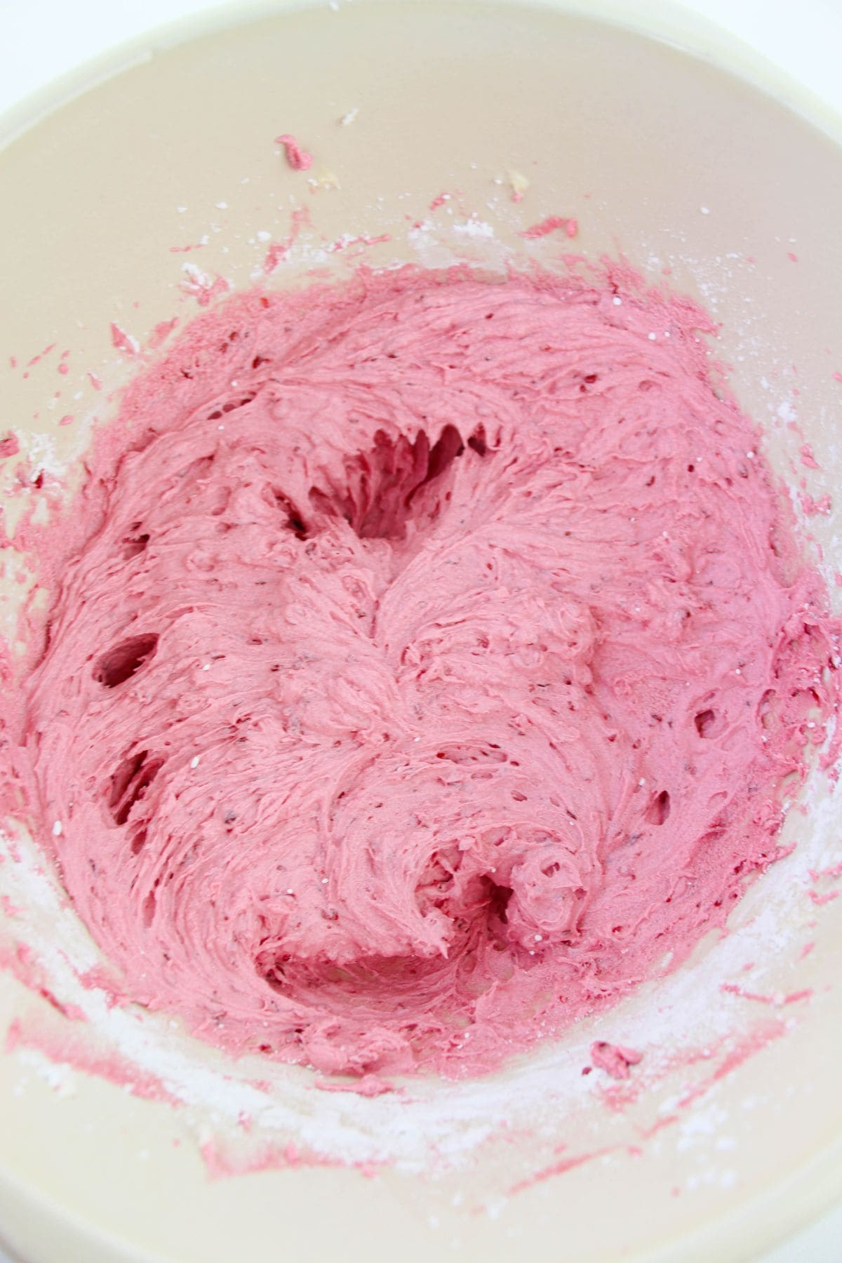 A bowl of vibrant pink raspberry buttercream is being mixed, with streaks of the creamy mixture and some flour visible around the edges.