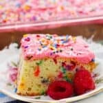 A slice of cake with pink raspberry buttercream frosting and colorful sprinkles sits on a floral plate with fresh raspberries, with the rest of the cake visible in the background.