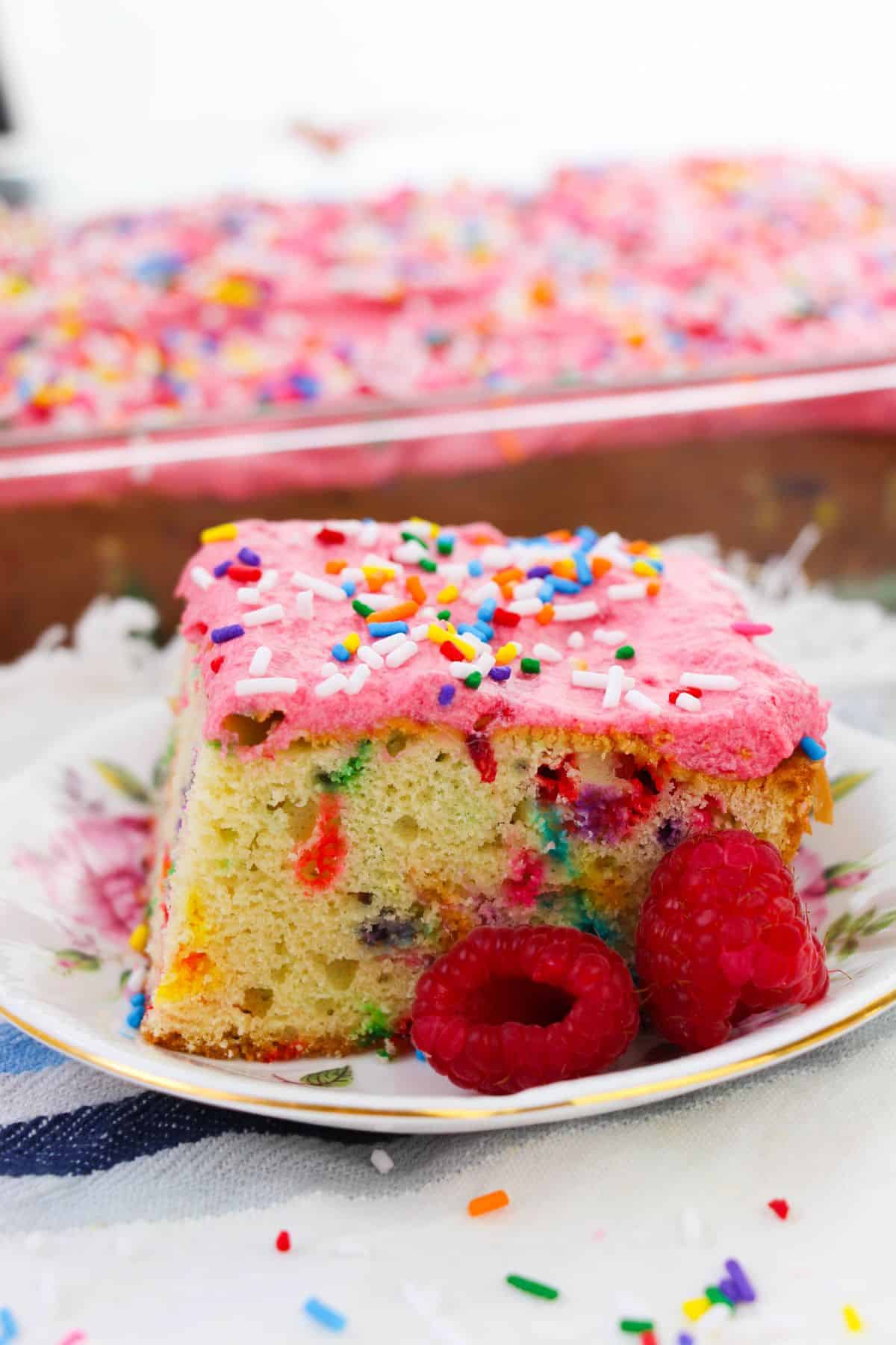 A slice of cake with pink raspberry buttercream frosting and colorful sprinkles sits on a floral plate with fresh raspberries, with the rest of the cake visible in the background.