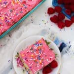 A slice of pink-frosted cake with rainbow sprinkles and raspberry buttercream sits on a plate with raspberries, next to a larger cake and a container of raspberries on a white cloth.