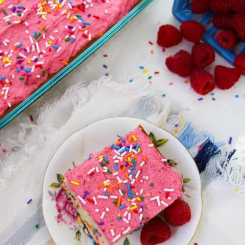 A slice of pink-frosted cake with rainbow sprinkles and raspberry buttercream sits on a plate with raspberries, next to a larger cake and a container of raspberries on a white cloth.