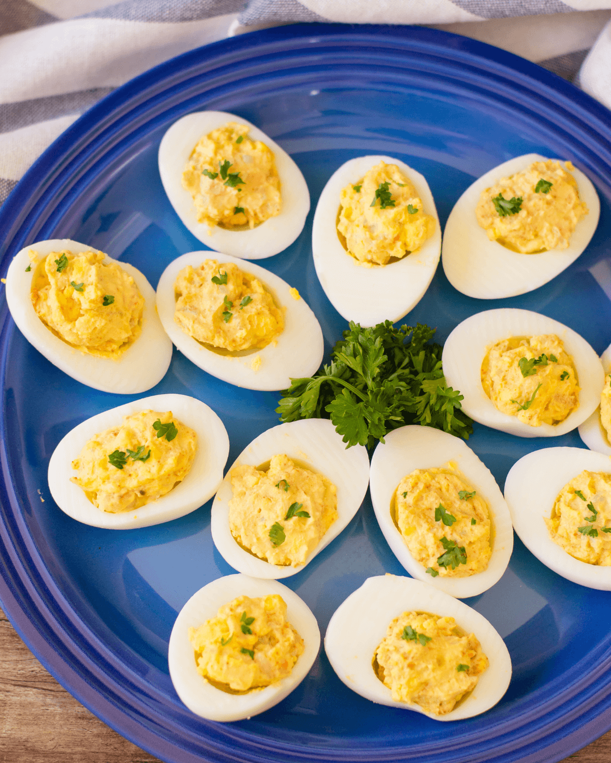 A blue plate with a dozen shrimp deviled eggs garnished with parsley on a wooden surface with a blue-striped cloth in the background.