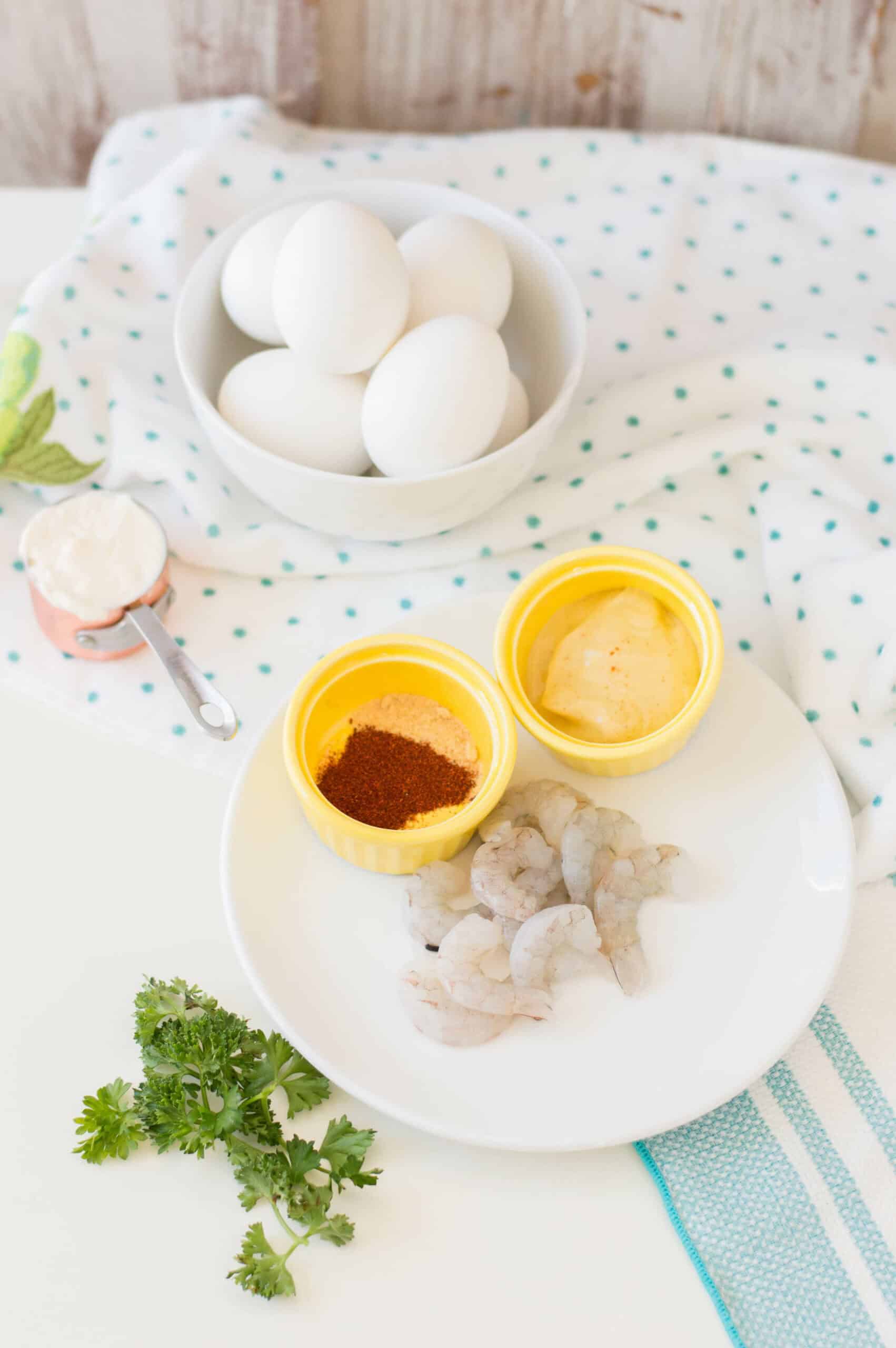 A kitchen counter with ingredients for cooking: a bowl of eggs, spices in small dishes, shrimp deviled eggs on a plate, and fresh herbs.