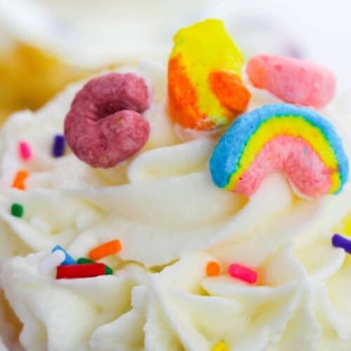 St. Patrick's Day cupcake with white frosting, crowned with colorful cereal pieces including a rainbow, and sprinkled with small rainbow sprinkles.