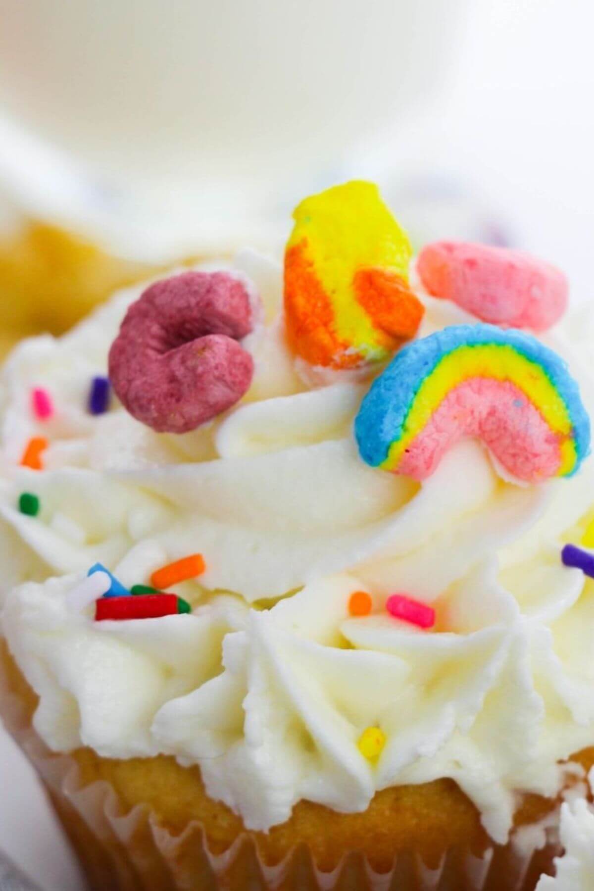 St. Patrick's Day cupcake with white frosting, crowned with colorful cereal pieces including a rainbow, and sprinkled with small rainbow sprinkles.