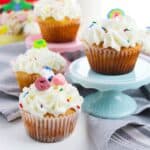 St. Patrick's Day cupcakes topped with white frosting and colorful candies sit on a table; one cupcake is on a blue stand. A cereal box and a glass of milk are in the background.