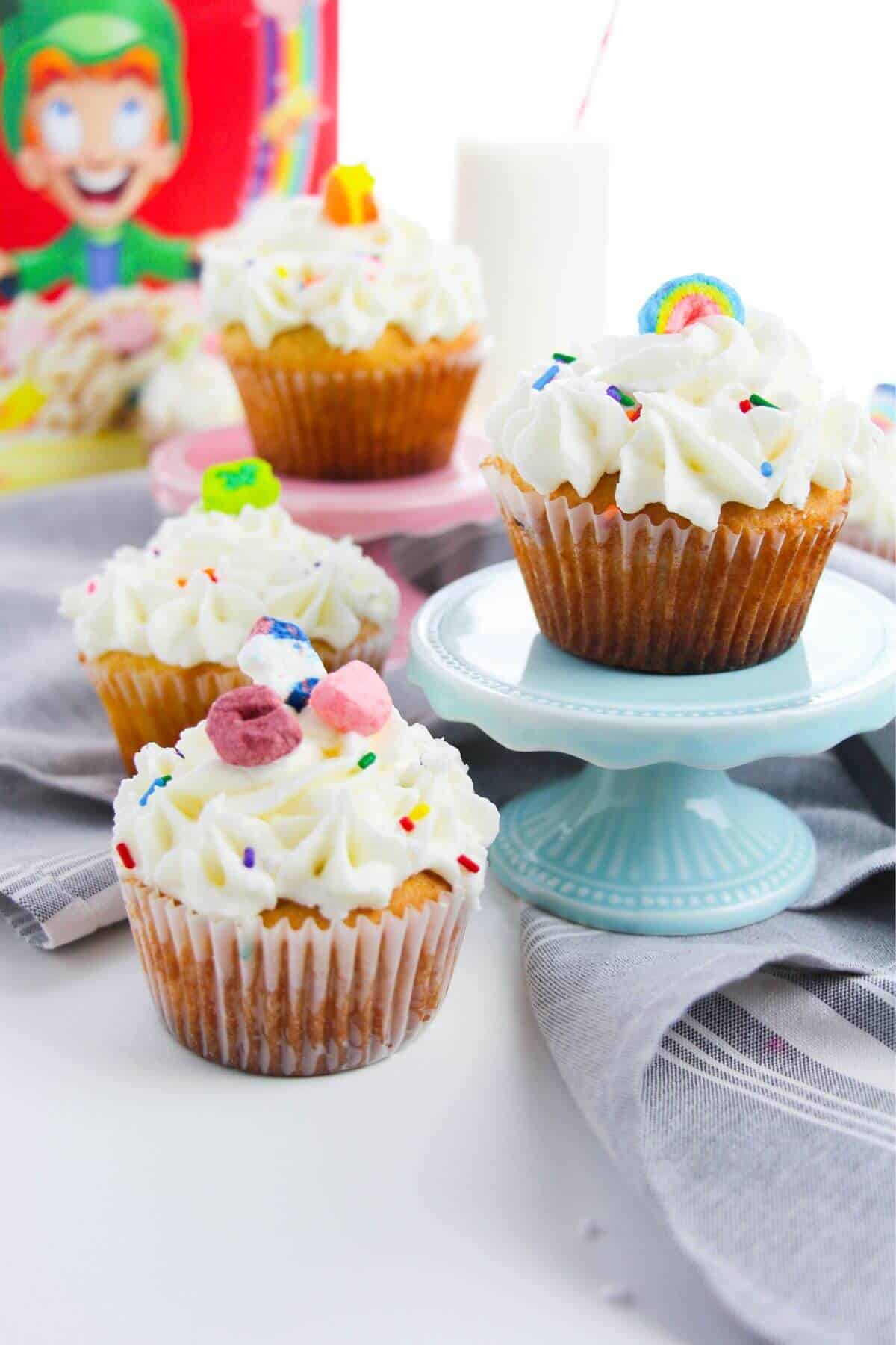 St. Patrick's Day cupcakes topped with white frosting and colorful candies sit on a table; one cupcake is on a blue stand. A cereal box and a glass of milk are in the background.