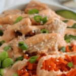 A bowl of food featuring diced chicken, green edamame, sliced cucumber, shredded carrots, sesame seeds, and a creamy teriyaki sauce drizzle.