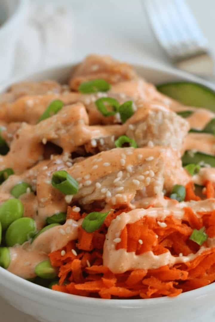 A bowl of food featuring diced chicken, green edamame, sliced cucumber, shredded carrots, sesame seeds, and a creamy teriyaki sauce drizzle.