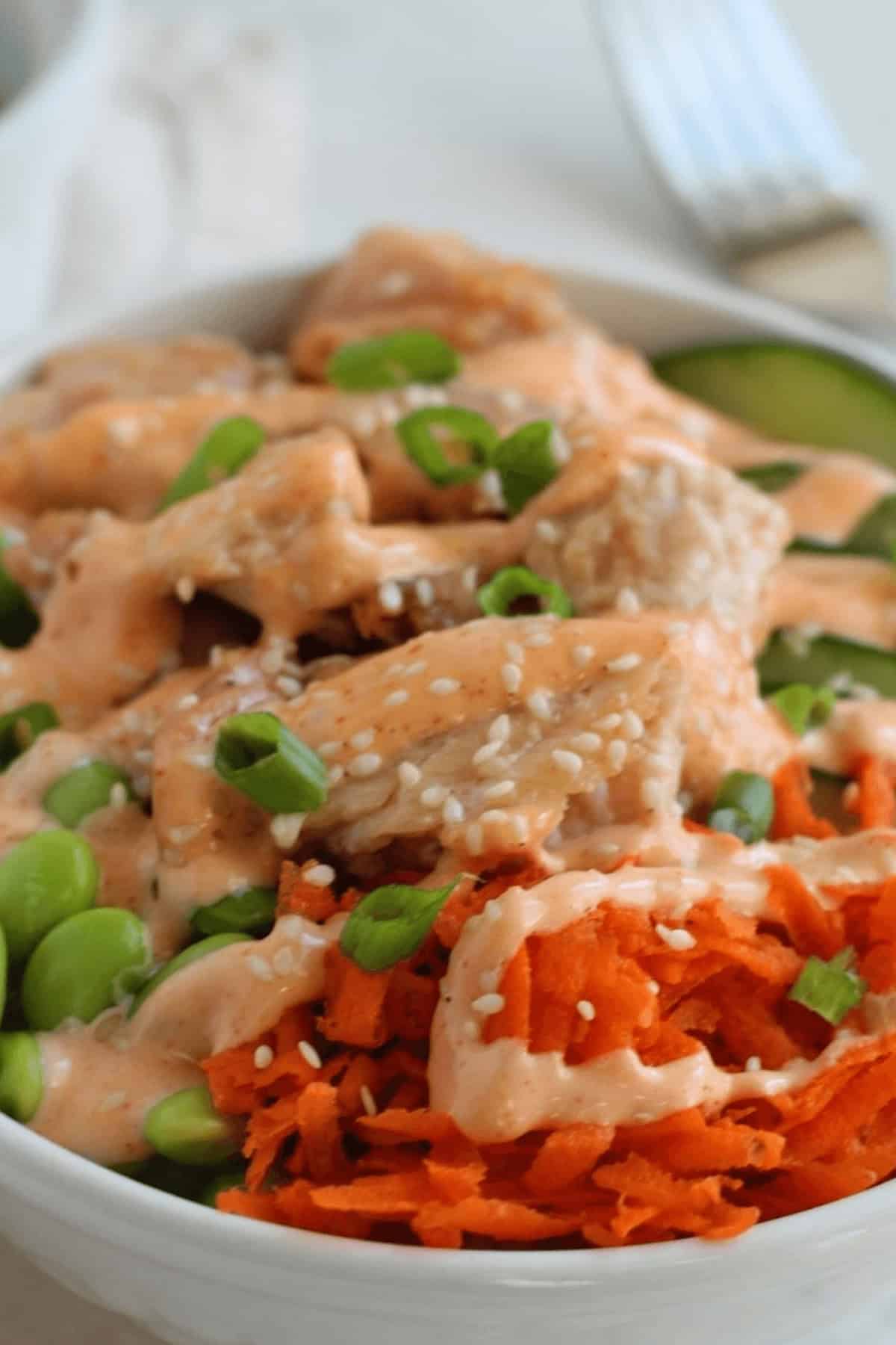 A bowl of food featuring diced chicken, green edamame, sliced cucumber, shredded carrots, sesame seeds, and a creamy teriyaki sauce drizzle.